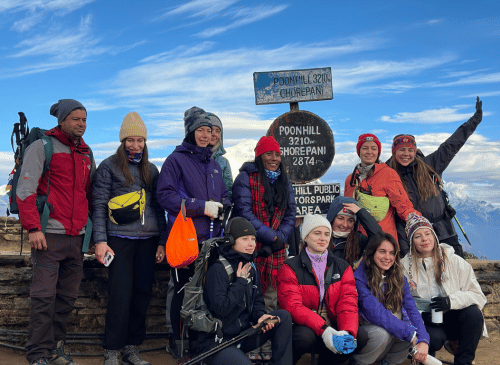 Group of international trekkers celebrating at the Poon Hill viewpoint sign (3210m/10531ft), showcasing the exhilarating short trek adventure included in the Nepal and Bhutan tour package.
