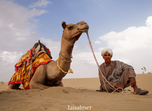 Two people (a couple) riding a camel during sunset on the golden sand dunes of the Thar Desert in Jaisalmer, Rajasthan. The sky is a warm orange and pink hue.